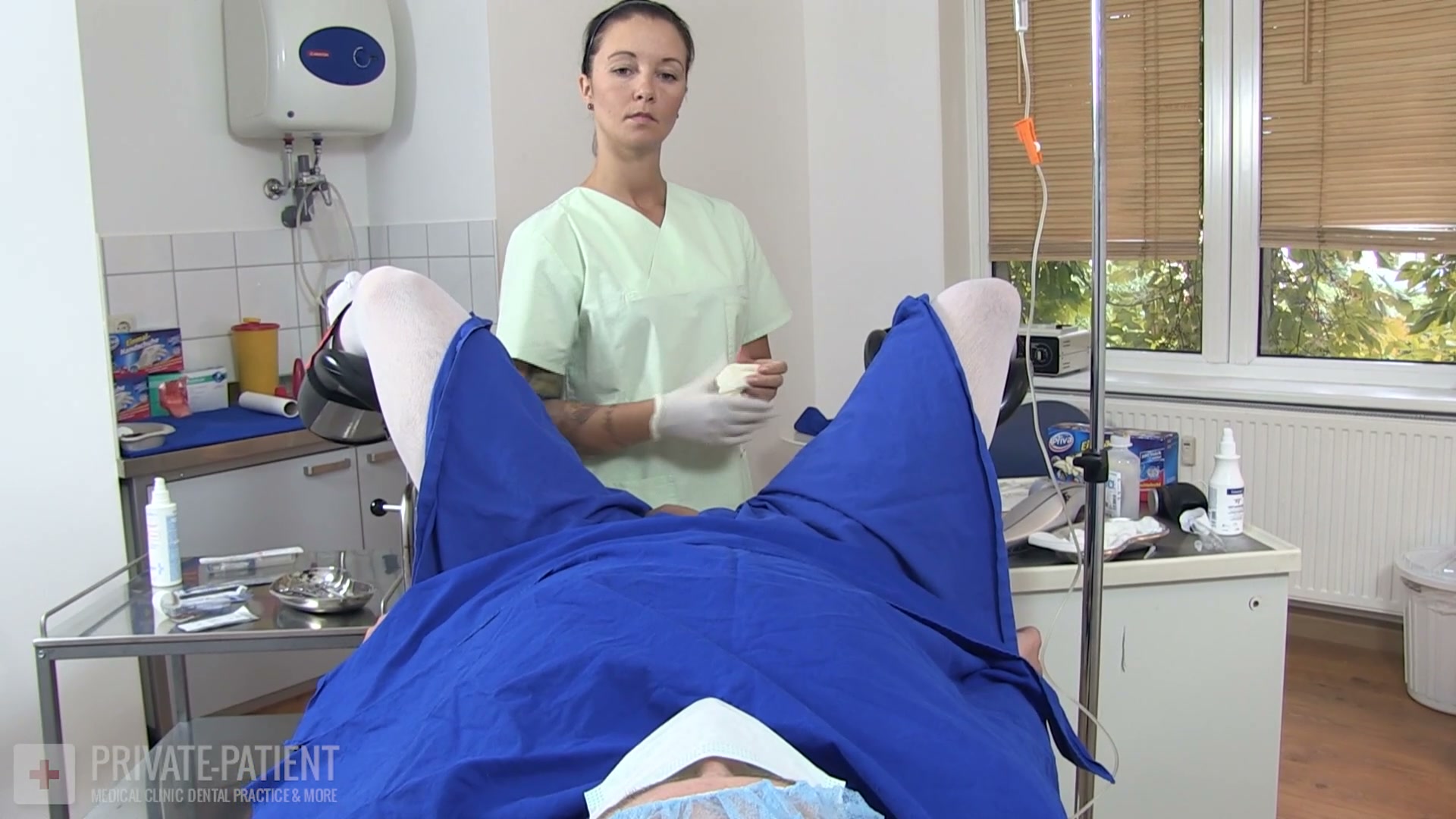 Dr. Eve gloved between the draped legs of a private patient in the free Ablatio Testis part two medfet video from Amara Fetish Clinic Belgium